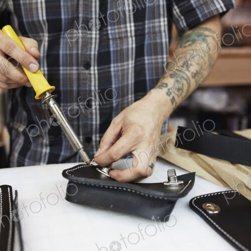 A leather worker and craftsman using a hand held tool on a blue leather bag on a workbench A leather worker and craftsman using a hand held tool on a blue leather bag on a workbench