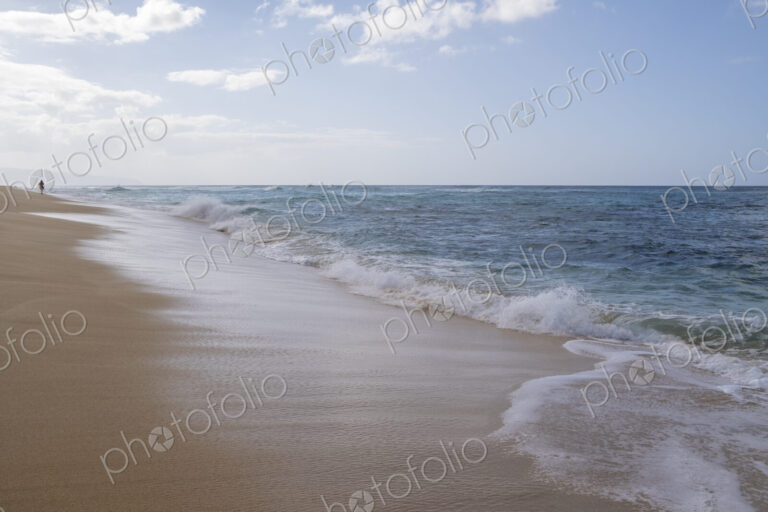 A lone person walks along Waimea Bay Beach, North Shore, waves breaking on the beach.