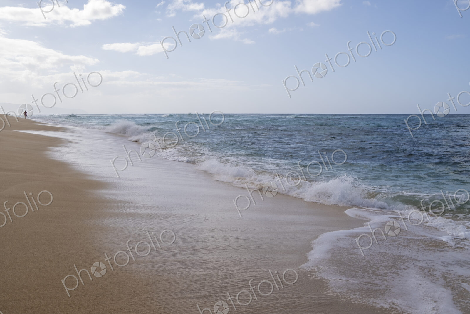 A lone person walks along Waimea Bay Beach, North Shore, waves breaking on the beach.
