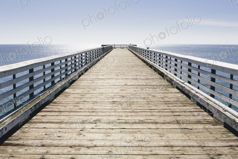 A long pier with railings extending out over the Pacific Ocean leading to the horizon.