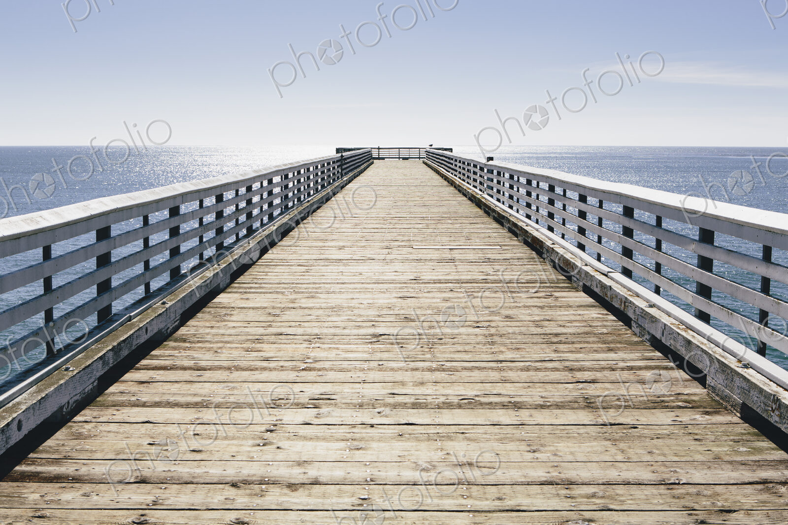A long pier with railings extending out over the Pacific Ocean leading to the horizon.