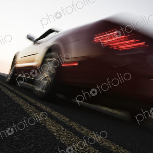 A low angle blurred view of a convertible sports car silhouetted on the road at sunset.