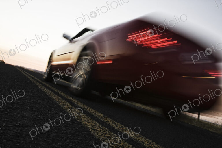 A low angle blurred view of a convertible sports car silhouetted on the road at sunset.