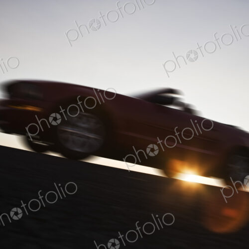 A low angle blurred view of a convertible sports car silhouetted on the road at sunset.