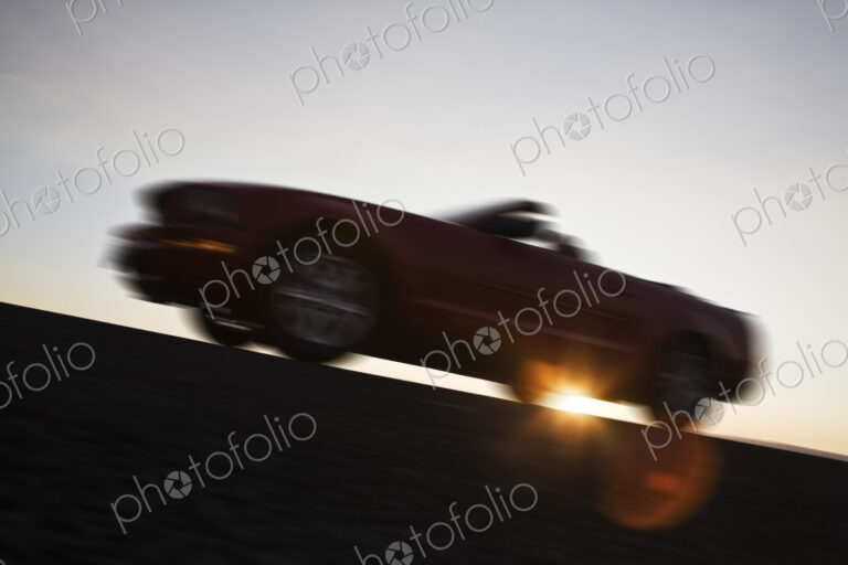 A low angle blurred view of a convertible sports car silhouetted on the road at sunset.