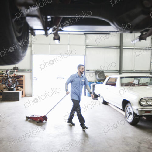 A male car mechanic pulls a portable car jack acrosss the floor of the car repair shop.