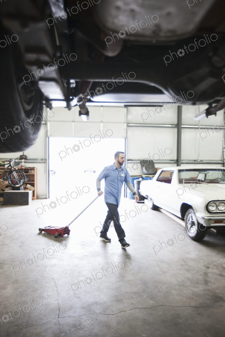 A male car mechanic pulls a portable car jack acrosss the floor of the car repair shop.