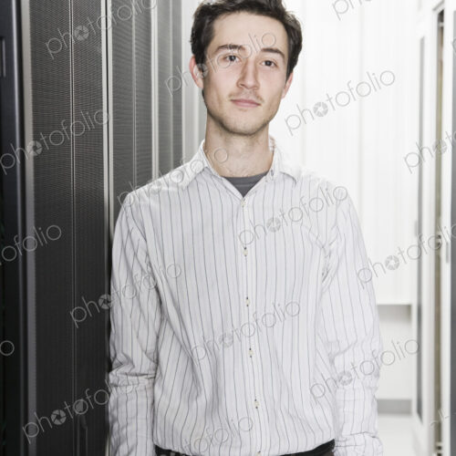 A male computer technician standing in an aisle of racks of servers in a computer…