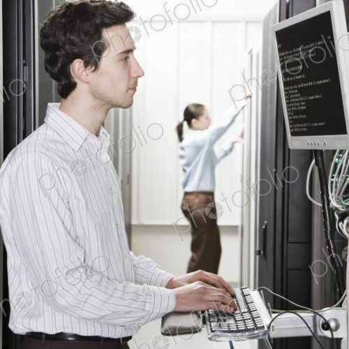 A male computer technician standing in an aisle of racks of servers in a computer…
