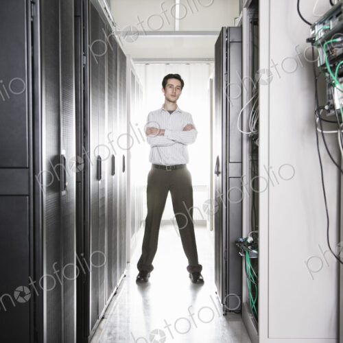 A male computer technician standing in an aisle of racks of servers in a computer…