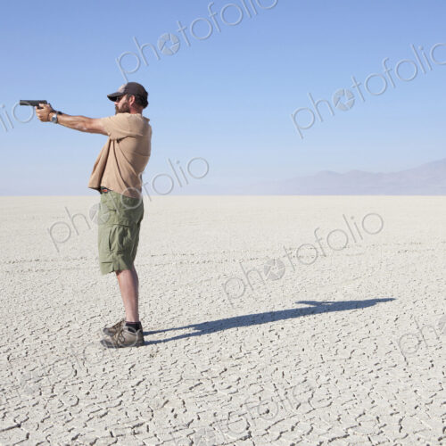 A man aiming a hand gun, holding it with his arm outstretched, standing in a vast,… A man aiming a hand gun, holding it with his arm outstretched, standing in a vast,…