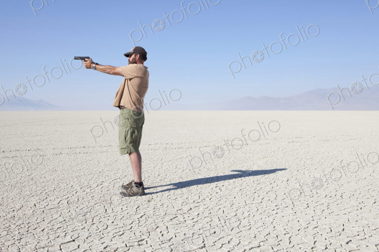 A man aiming a hand gun, holding it with his arm outstretched, standing in a vast,…