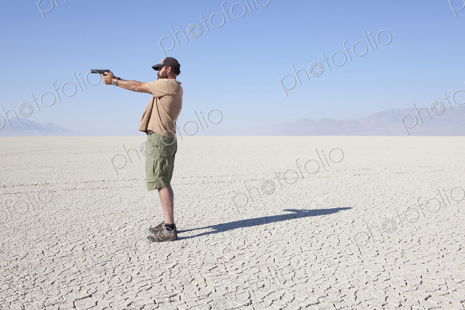 A man aiming a hand gun, holding it with his arm outstretched, standing in a vast,…
