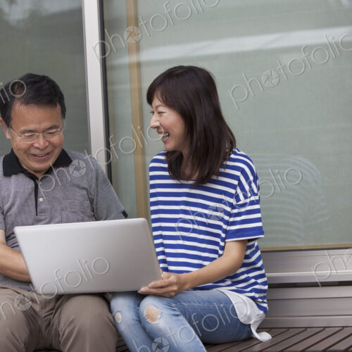 A man and a women sitting outside a house. Holding a laptop computer.