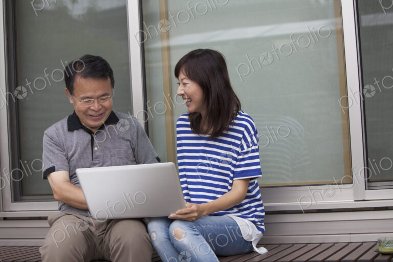 A man and a women sitting outside a house. Holding a laptop computer.