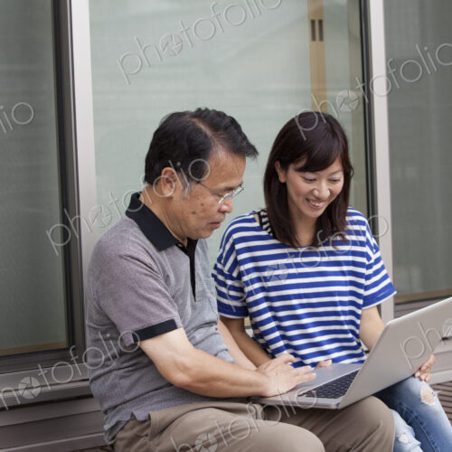 A man and a women sitting outside a house. Holding a laptop computer.