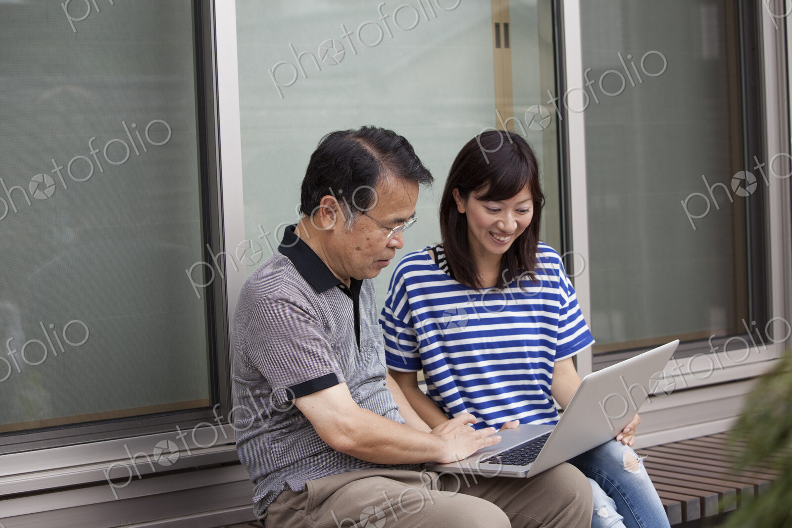 A man and a women sitting outside a house. Holding a laptop computer.