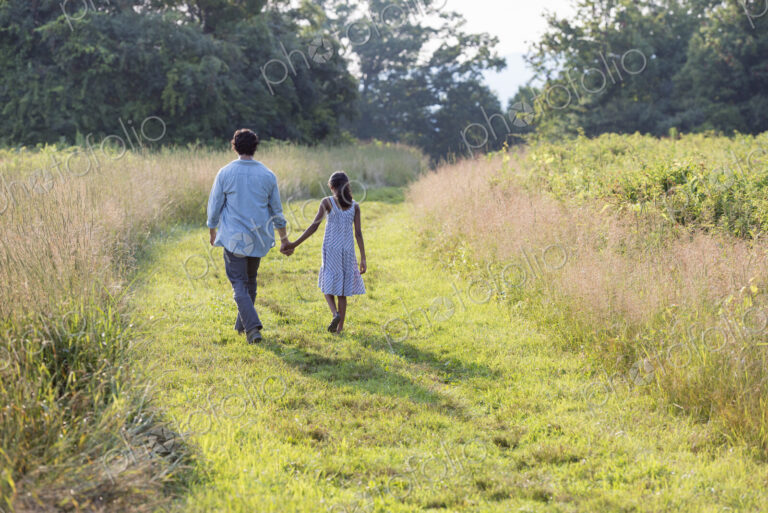 A man and a young girl walking down a mown path in the long grass holding hands.