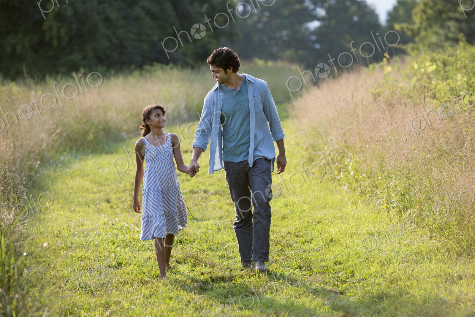A man and a young girl walking down a mown path in the long grass holding hands.