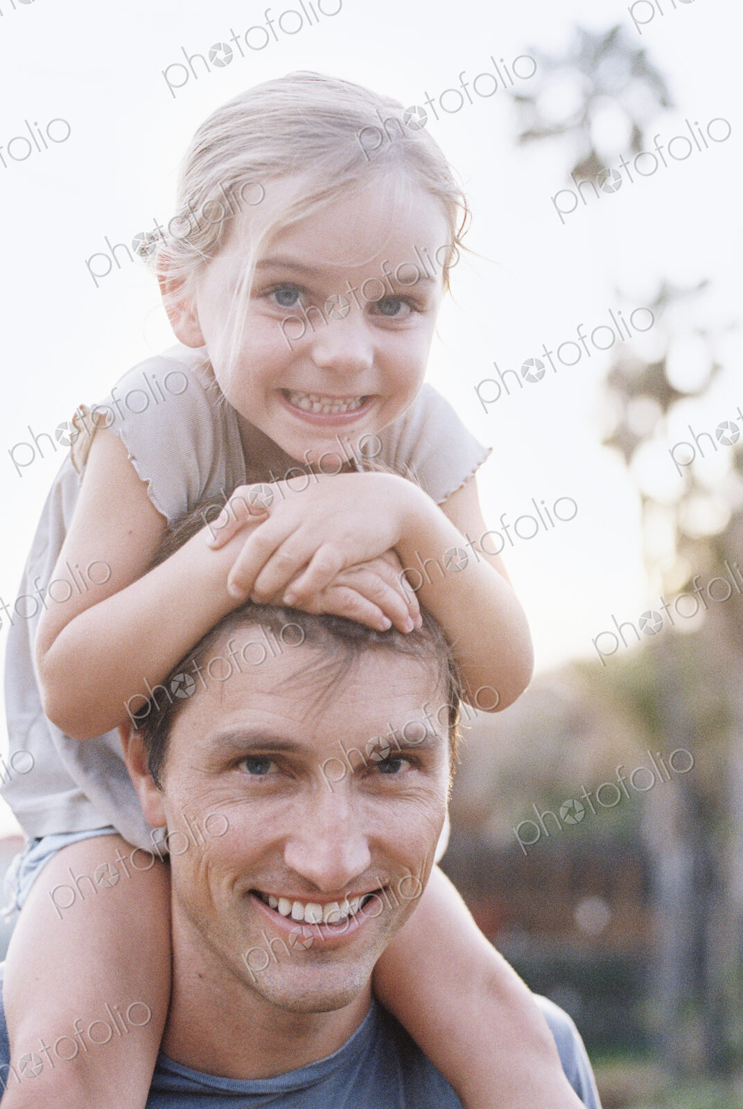 A man and his daughter smiling at the camera, the child sitting on his shoulders.