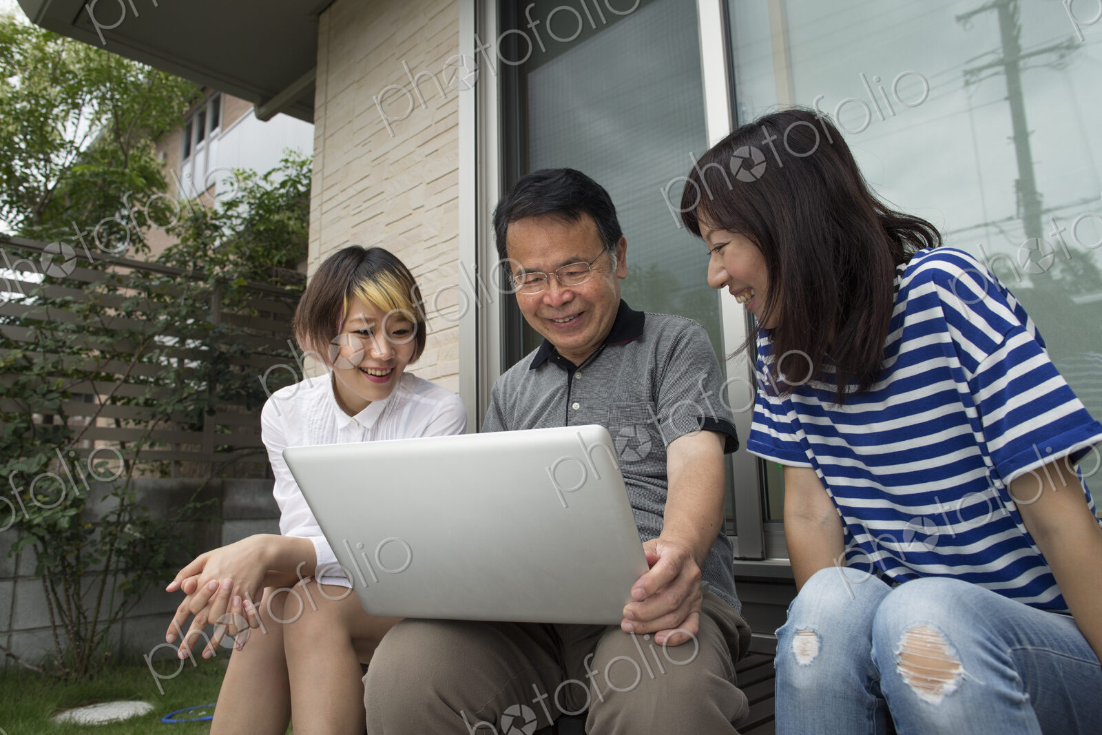 A man and two women sitting outside a house. Holding a laptop computer.