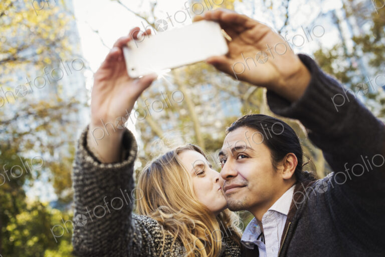 A man and woman kissing and talking a selfy outdoors in a city.