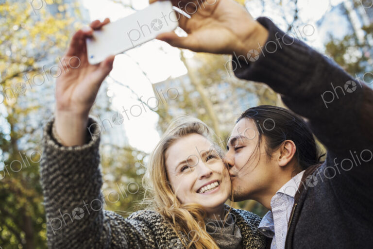 A man and woman kissing and talking a selfy outdoors in a city.