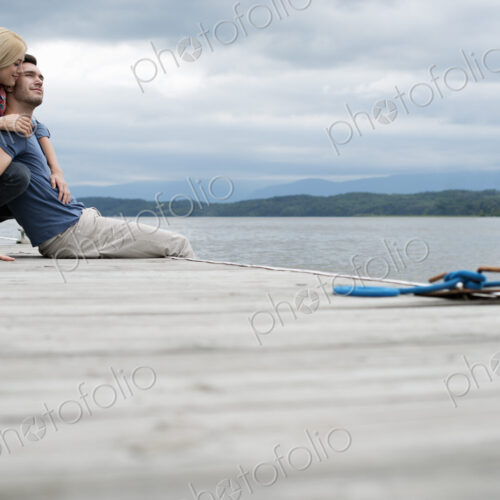 A man and woman seated on a jetty by a lake.