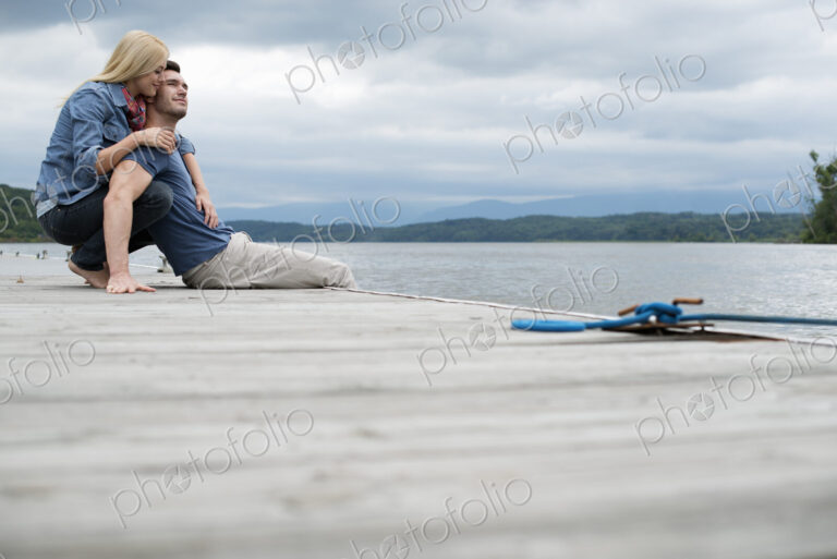 A man and woman seated on a jetty by a lake.