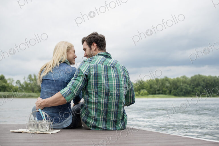 A man and woman sitting close together on a jetty by a lake.