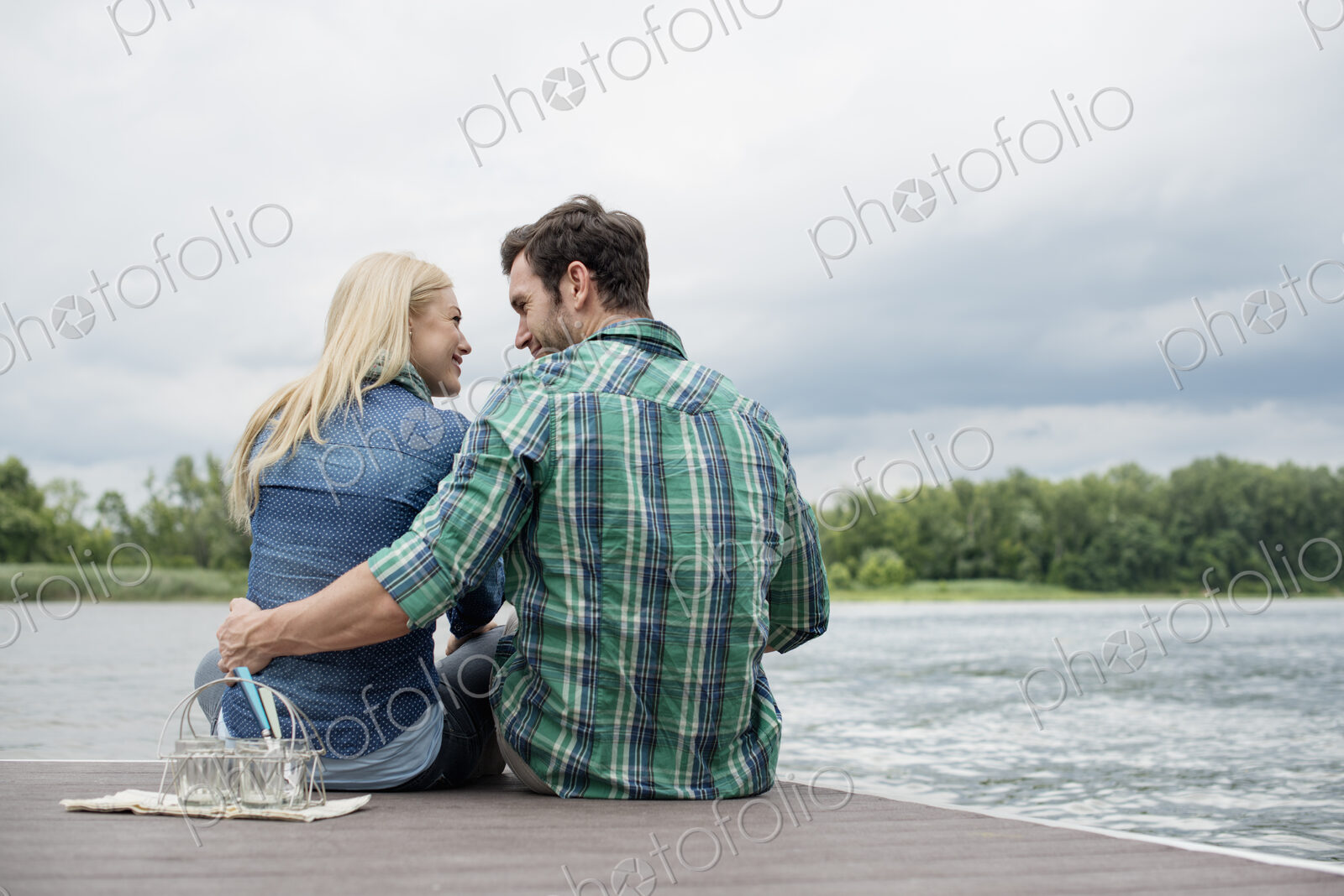 A man and woman sitting close together on a jetty by a lake.