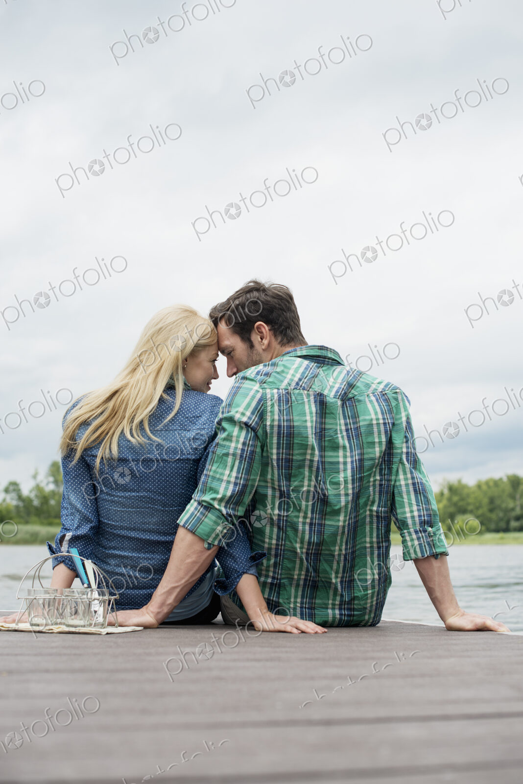 A man and woman sitting close together on a jetty by a lake.