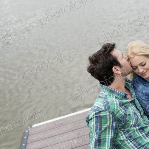 A man and woman sitting close together on a jetty by a lake.