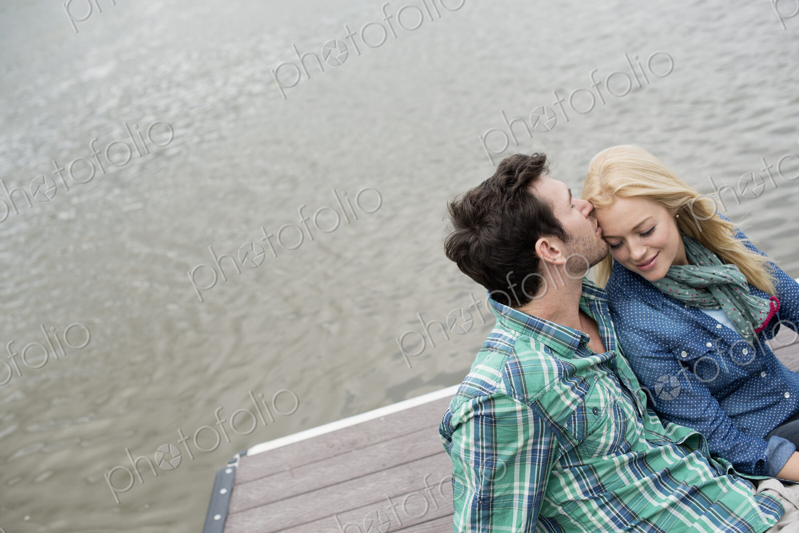 A man and woman sitting close together on a jetty by a lake.