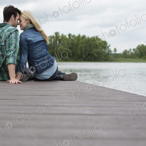 A man and woman sitting close together on a jetty by a lake.