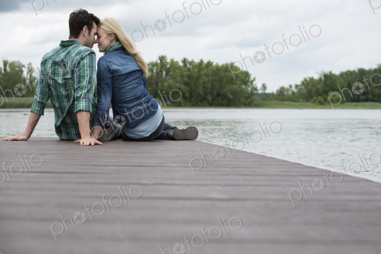 A man and woman sitting close together on a jetty by a lake.