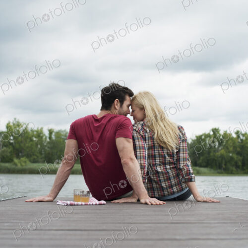 A man and woman sitting close together on a jetty by a lake.