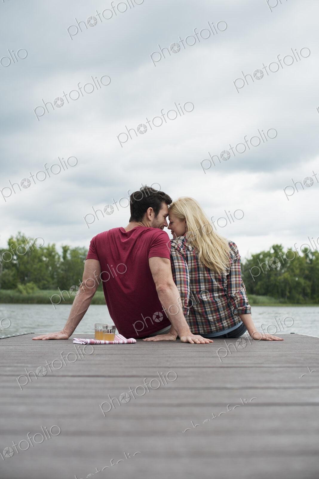 A man and woman sitting close together on a jetty by a lake.