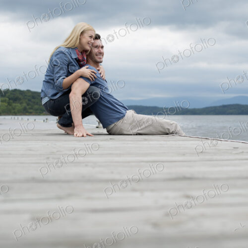 A man and woman sitting close together on a jetty by a lake.