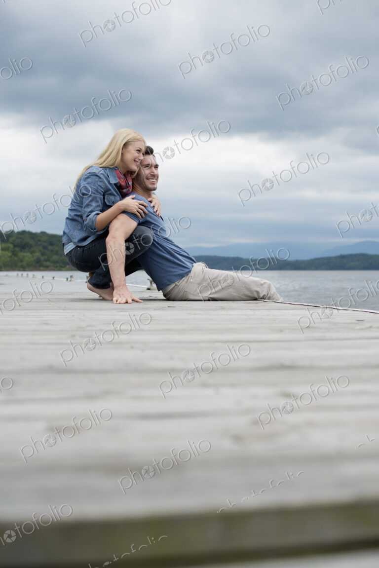 A man and woman sitting close together on a jetty by a lake.
