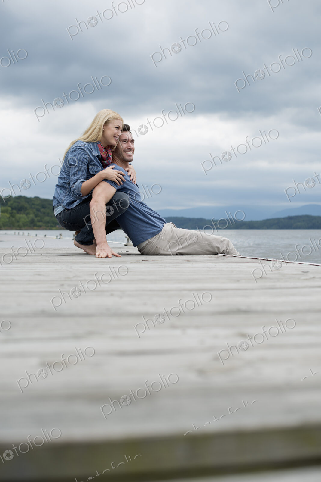 A man and woman sitting close together on a jetty by a lake.