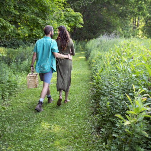 A man and woman walking through a meadow, through the long grass, carrying a picnic…