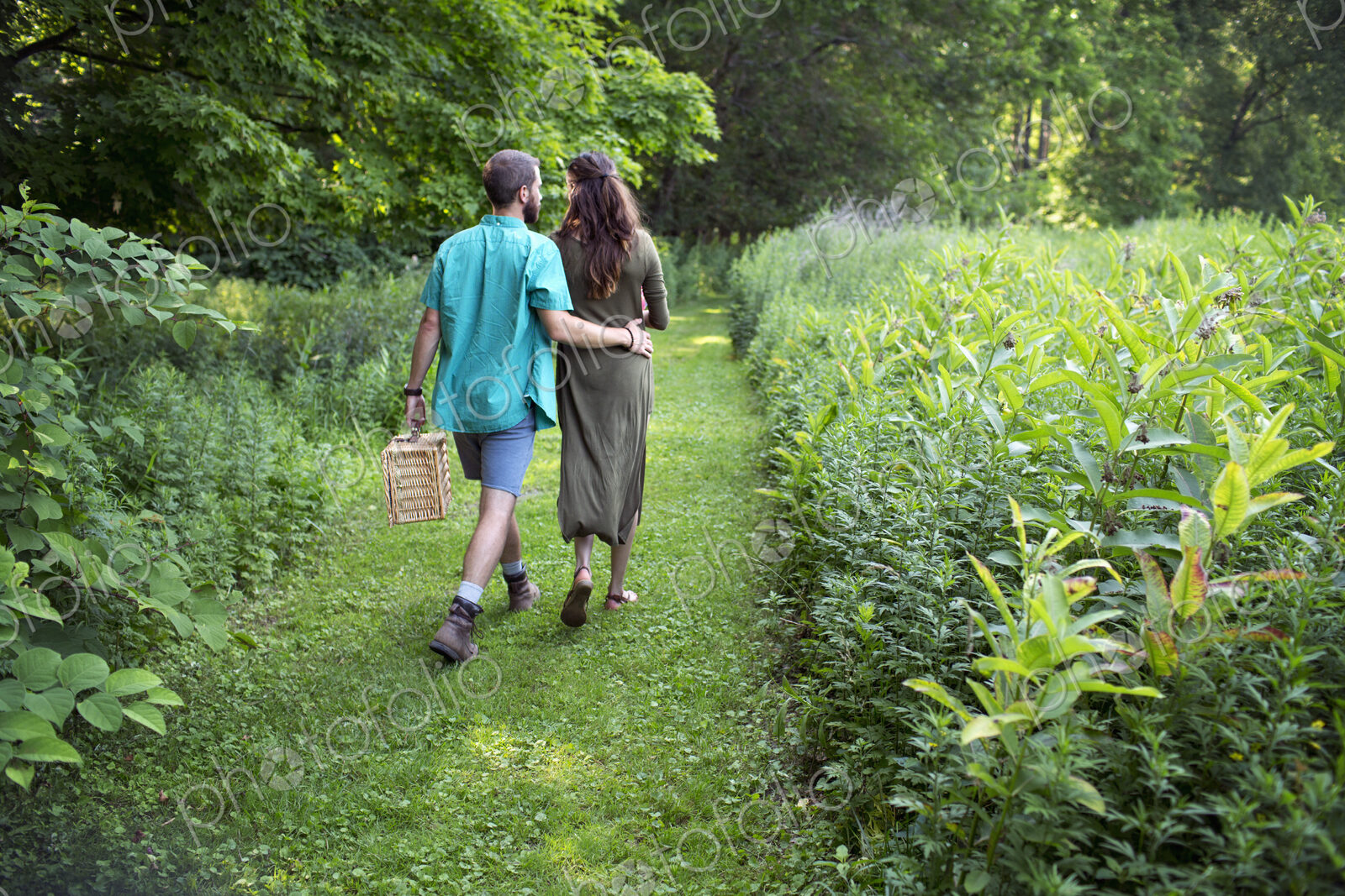 A man and woman walking through a meadow, through the long grass, carrying a picnic…