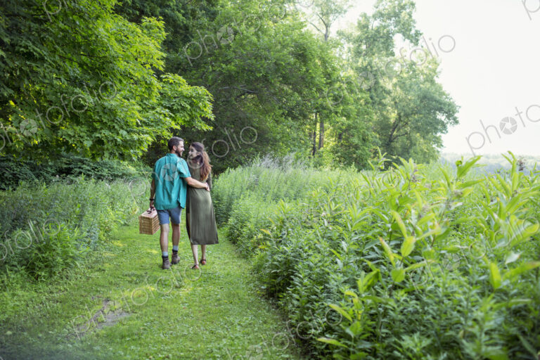 A man and woman walking through a meadow, through the long grass, carrying a picnic…