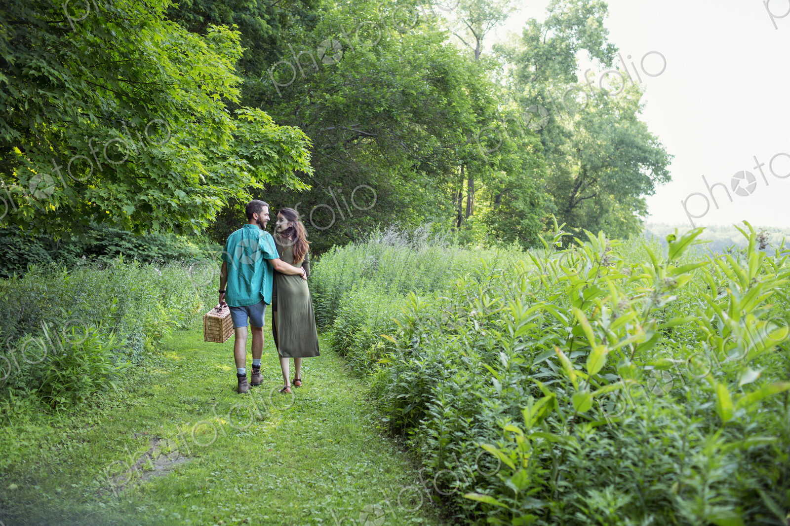 A man and woman walking through a meadow, through the long grass, carrying a picnic…