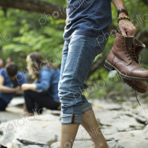 A man carrying his boots walking on the rocks by a river.