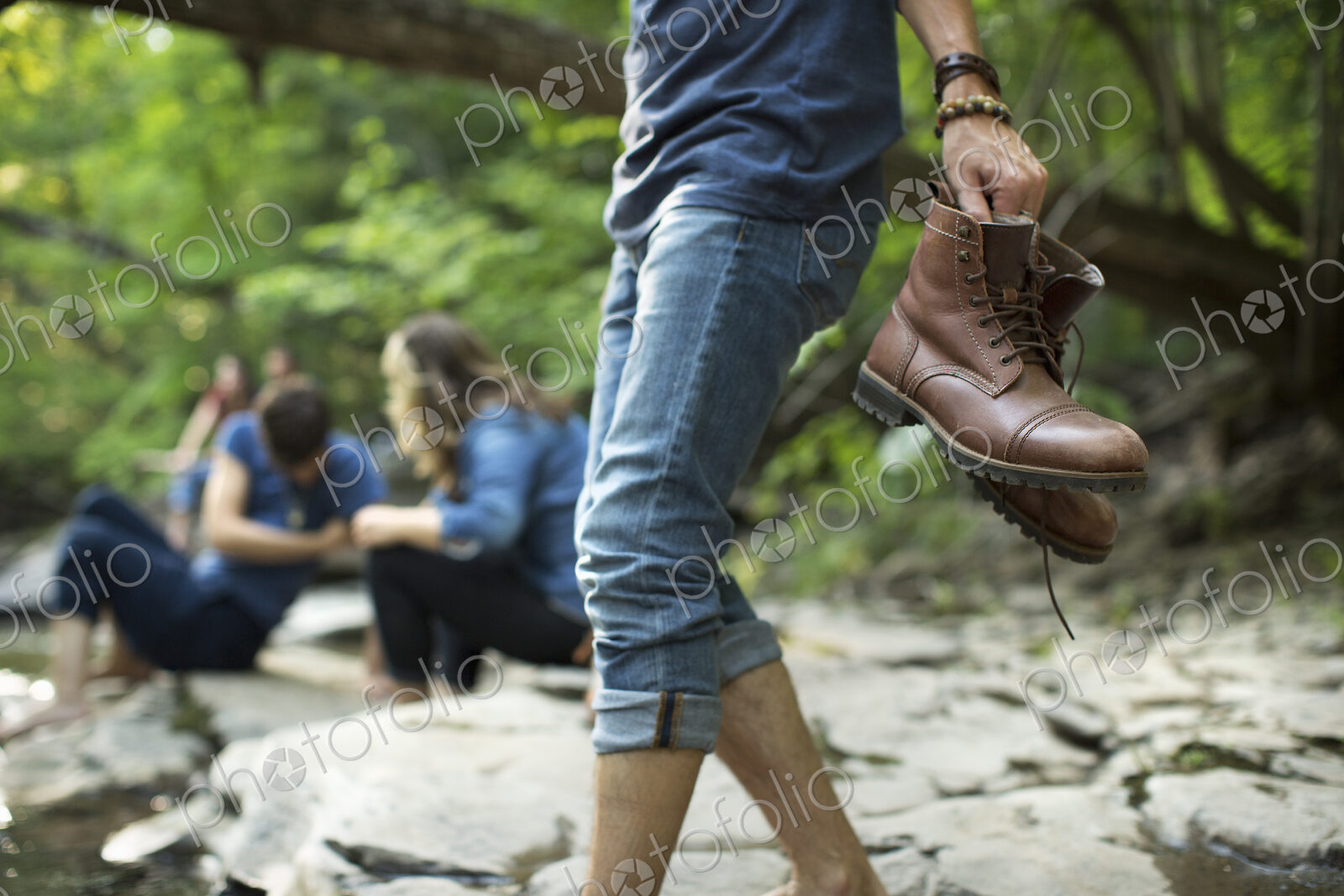 A man carrying his boots walking on the rocks by a river.