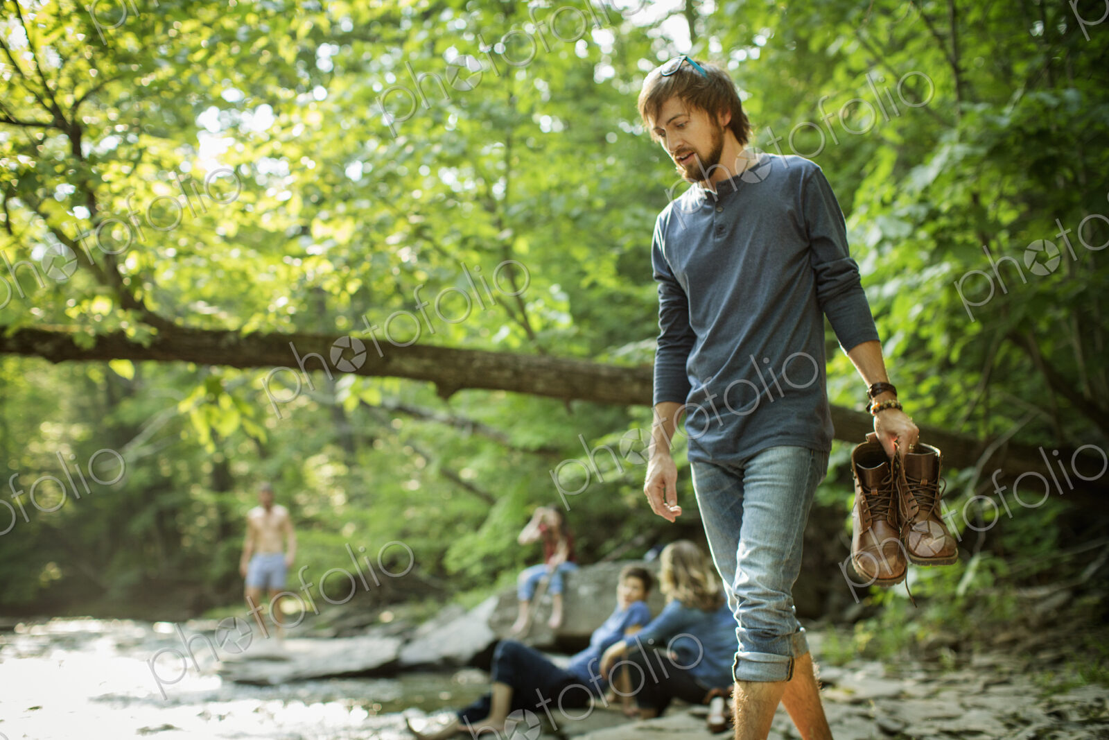 A man carrying his boots walking on the rocks by a river.