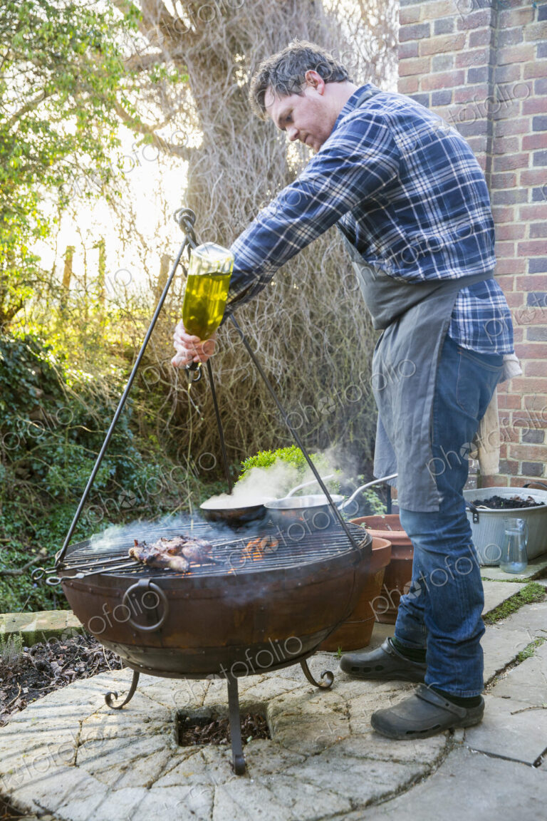 A man cooking roasting game birds over an open fire, pouring olive oil from a bottle…