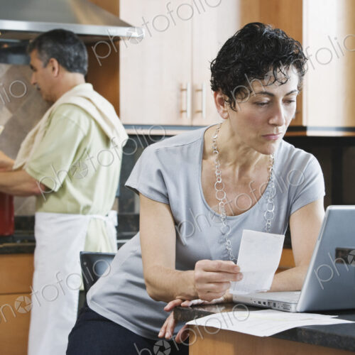 A man cooking supper at the hob, and a woman using a laptop and checking paperwork in… A man cooking supper at the hob, and a woman using a laptop and checking paperwork in…